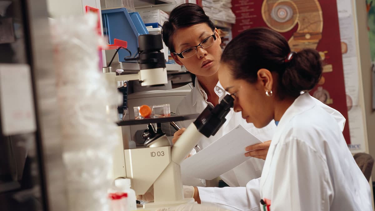 Two women in lab coats looking through a microscope.