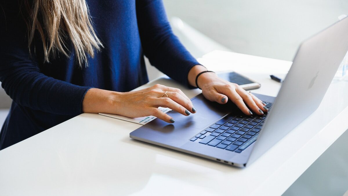 A woman in blue long sleeve shirt typing on a laptop at a desk.