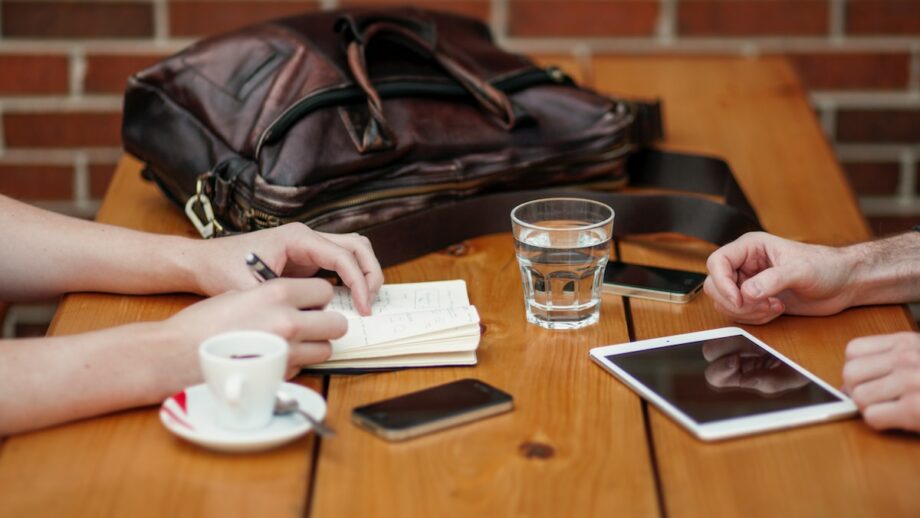 Two people sitting at a table with a tablet and a smartphone writing on a notebook.