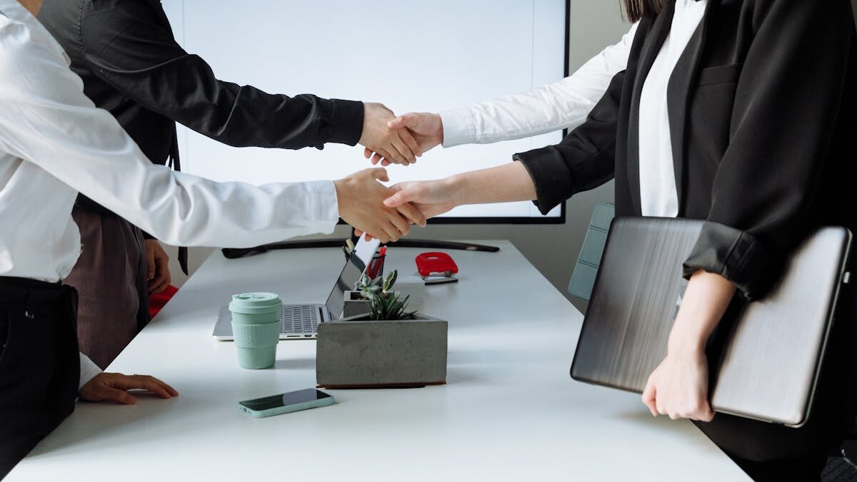 Four individuals shaking hands over a table in an office environment, suggesting a professional agreement or greeting.