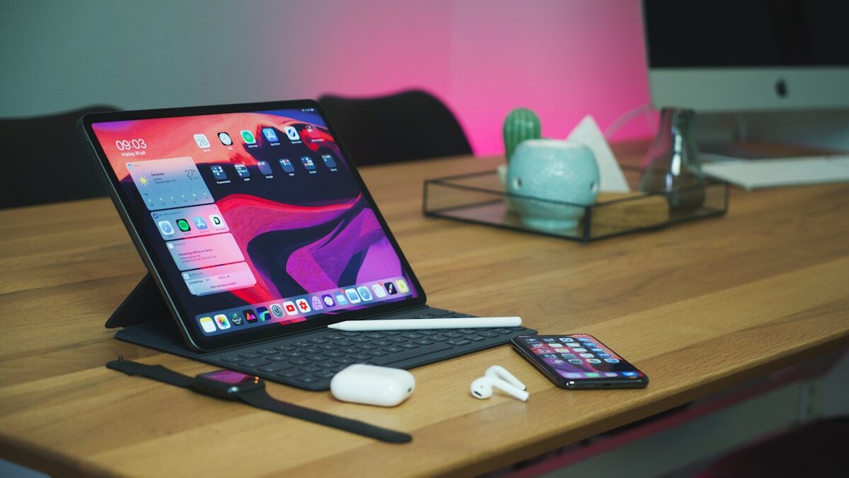 A modern workspace featuring an ipad with a keyboard on a stand, an iphone, apple pencil, airpods, and a desktop computer in the background, all arranged neatly on a desk with ambient lighting.