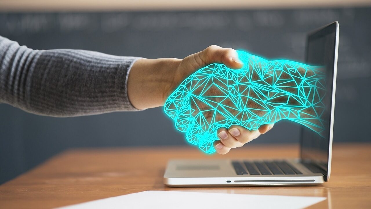A person's hand extends towards a laptop screen with a graphic of a blue, geometric handshake appearing, symbolizing digital connectivity or online agreements.