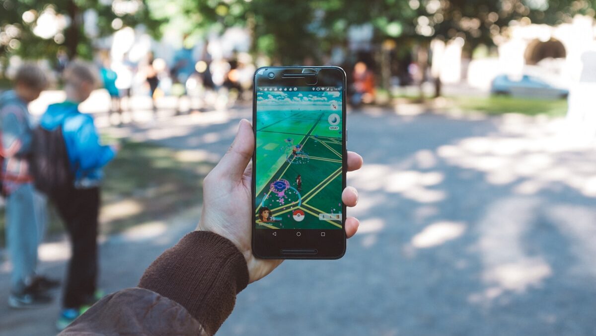A person's hand holding a smartphone displaying a location-based augmented reality game, with blurred figures and trees in the background in a park setting.