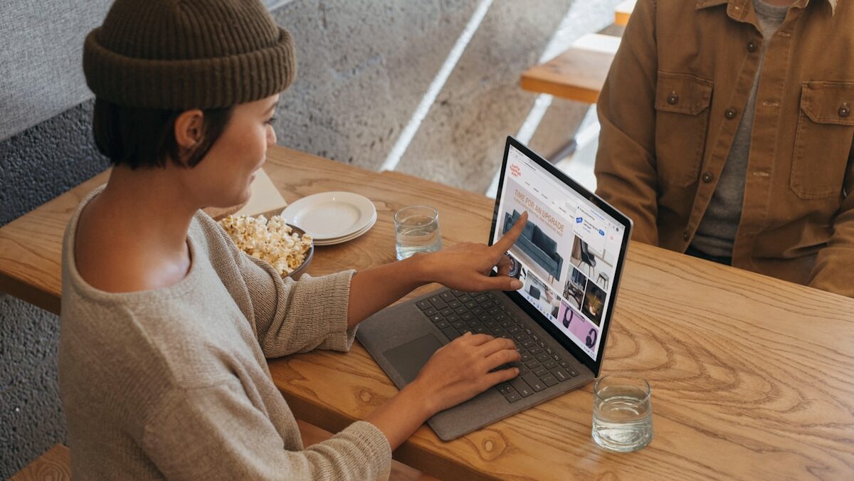 A woman in a beanie uses a laptop at a wooden table, pointing at the screen to show something to a person sitting across from her. they have plates and a glass of water on the table.